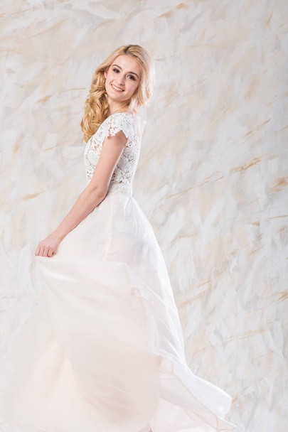 fashionable wedding dress, beautiful blonde model, bride hairstyle and makeup concept - happy young woman in long luxury white gown indoors on light background, stunning female posing with a smile - Foto, afbeelding