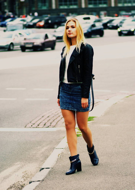 ein blondes Mädchen mit langen Haaren in Jeansrock und Ledermantel steht auf einer Straße. - Foto, Bild