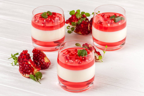 Romig vanille panna cotta met rode gelei in mooie glazen, verse rijpe granaatappel op witte houten achtergrond. - Foto, afbeelding