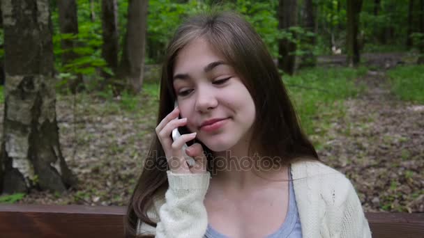 Young attractive girl speak to phone on bench. Summer park. Smile. HD video footage shooting static camera. - Felvétel, videó