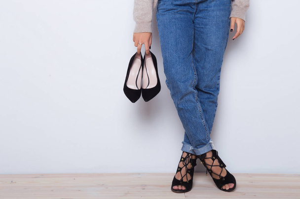 Choice which high heel shoes wear today. Fashion jeans on a girl, girl holding a pair of shoes. Thin legs of fashion girl. Shapely female legs in high heel shoes and jeans near a white wall  - Фото, изображение