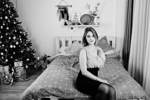 Blonde girl in dress posed on bed near new year tree with christ - 写真・画像