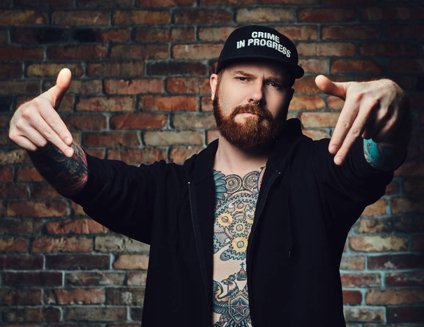 Portrait of redhead bearded male with tattoos on arms and chest, dressed in black sports jacket over the wall of a brick. - Photo, Image
