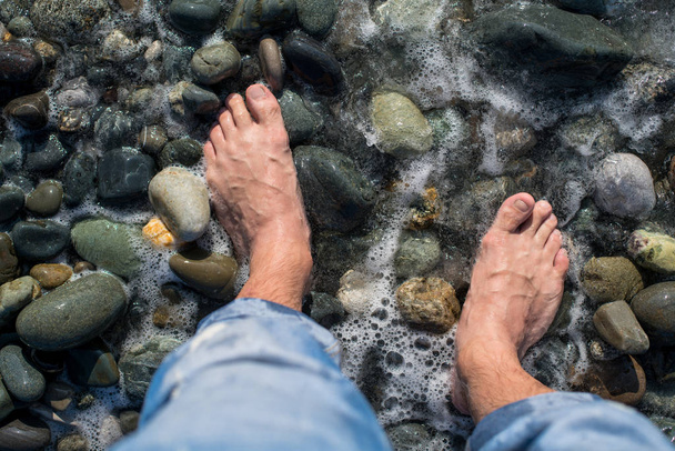 Bare feet of a man who stands on a pebble beach next to the sea wave. Water foams. - Photo, image