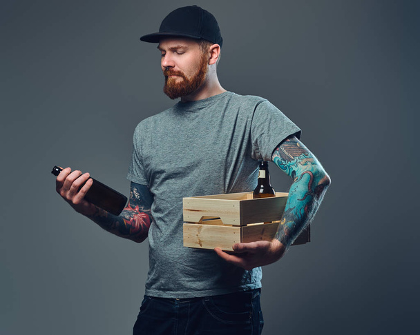 彼の腕に入れ墨をしたキャップでひげを生やした男性の肖像画を保持しているビール瓶の木箱. - 写真・画像