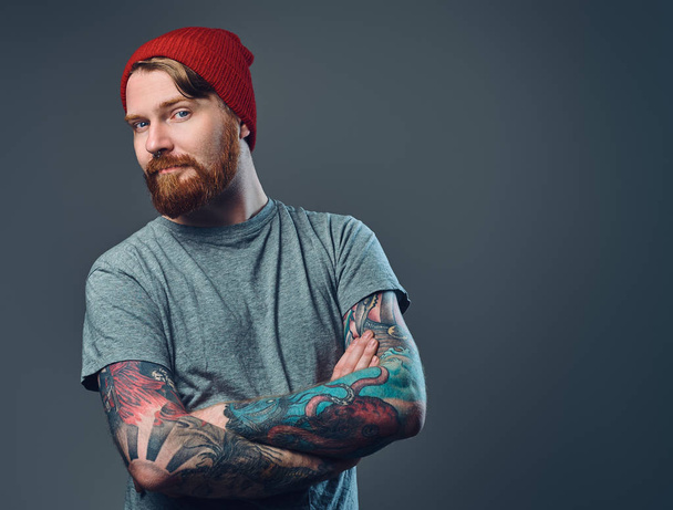 Vörös hajú, szakállas férfi, a tetoválás a karját, pózol, mint szürke háttér. - Fotó, kép
