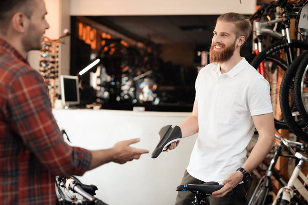 A seller at a bicycle store helps a young buyer choose a saddle for a bicycle. A man with a beard shows the client different types of goods. They both have a good mood. - Foto, imagen