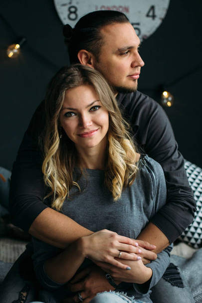 Boyfriend and girfriend touching each other sitting on bed with grey blankets and pillows. Cozy room with lights, romantic and love concept. Couple kissing and smiling - Фото, зображення