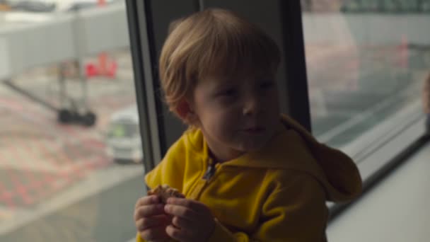 Slowmotion shot of a young boy in a yellow jacket eating a cookie sittting in front of a big window at an airport - Footage, Video