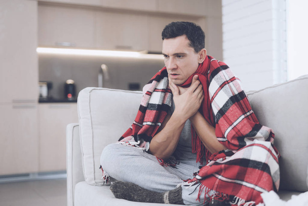 A man with a cold sits on the couch, hiding behind a red rug. He has a sore throat - Foto, Imagen