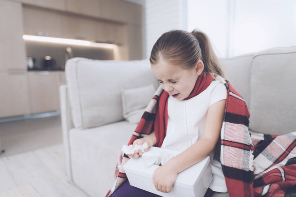 Little sick girl sits on a white couch wrapped in a red scarf. She sneezes, taking out a napkin - Photo, Image