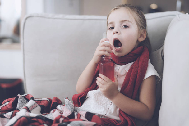Little sick girl sits on a white couch wrapped in a red scarf. She splashes her throat with a medicinal spray - Fotoğraf, Görsel