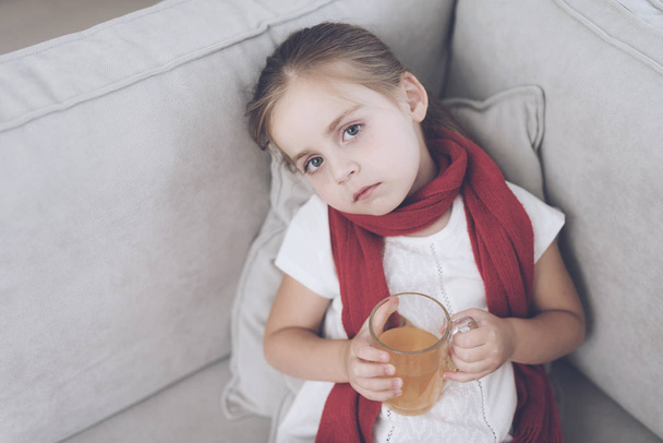 Little sick girl sits on a white couch wrapped in a red scarf. She is sitting with a cup of medicinal tea - Foto, Imagem