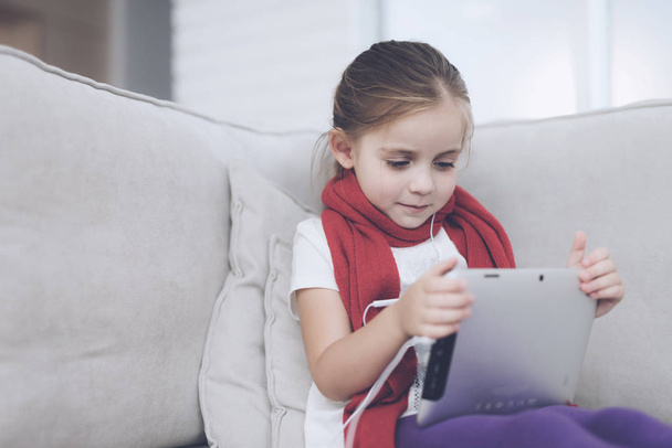 Little sick girl sits on a white couch wrapped in a red scarf. She holds a gray tablet in her hands - Foto, afbeelding