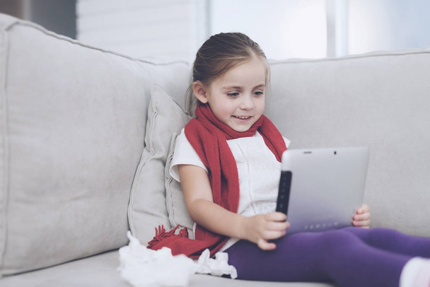 Little sick girl sits on a white couch wrapped in a red scarf. She holds a gray tablet in her hands - Foto, imagen