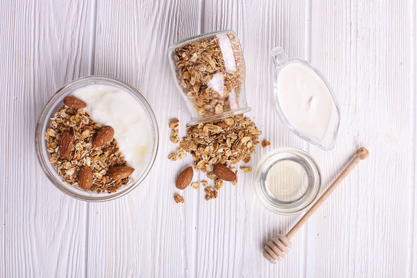 Granola superfood with almond and cashew nuts, dry fruits, raisins cherry in the glass jar on the white wooden table, top view, bright light. Healthy breakfast granola with greek yogurt, honey, mix nuts on white wooden background top view, copy space - Foto, immagini