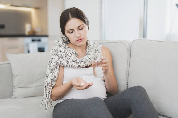 Sick pregnant woman sitting at home on the couch. She's getting ready to drink a pill. - Photo, Image