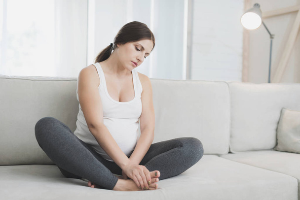 Eine schwangere Frau sitzt zu Hause auf einem hellen Sofa. Ihre Beine schmerzen - Foto, Bild