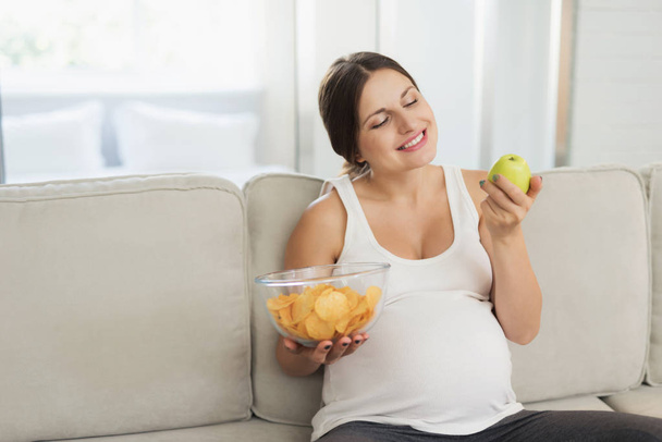 A pregnant woman is sitting on a light sofa at home. She holds a vase of chips in her hand and eats an apple - 写真・画像