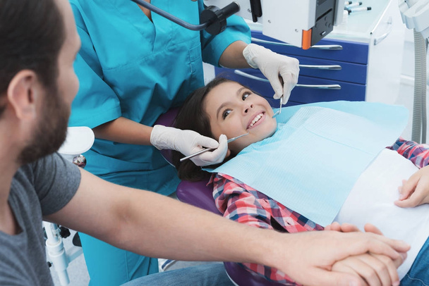 Une dentiste soigne une fille. Elle est assise sur la chaise dentaire. Elle a peur et son père lui tient la main
. - Photo, image