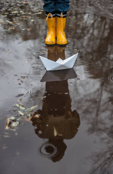 Child with Yellow Rain Boots and a little White Paper boat / Ship: Playing in a puddle, imagining his adventures - Photo, Image
