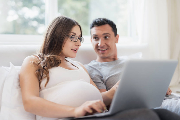 A pregnant woman and a man are lying on the bed. A woman is holding a laptop on her lap, a man is holding a smartphone. - 写真・画像
