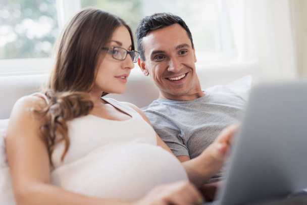 A pregnant woman and a man are lying on the bed. A woman is holding a laptop on her lap, a man is holding a smartphone. - Foto, imagen