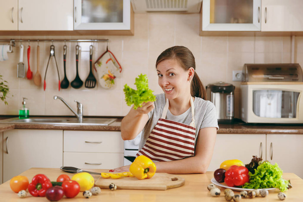 eine junge attraktive Frau in einer Schürze mit Salatblatt in der Küche. Diätkonzept. Gesunder Lebensstil. Kochen zu Hause. Essen zubereiten. - Foto, Bild