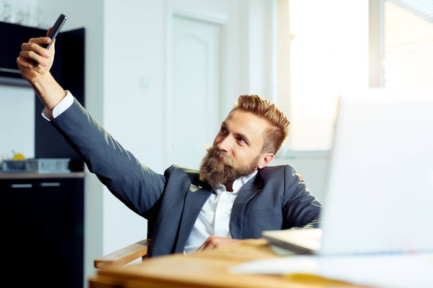 Happy businessman sitting at the table and making selfie photo on smartphone in office - Fotografie, Obrázek