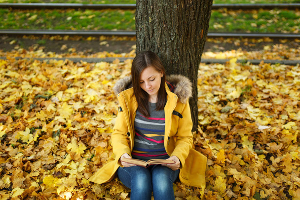 A beautiful happy smiling brown-haired woman in yellow coat and jeans sitting under the maple tree with a red book in fall city park on a warm day. Autumn golden leaves. Reading concept - Photo, Image