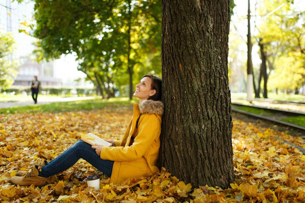 Kaunis iloinen ruskeahiuksinen nainen keltaisessa takissa ja farkut istuu vaahteran puun alla kupin teetä tai kahvia ja punainen kirja syksyllä kaupungin puistossa. Syksyn kultaisia lehtiä. Lukukäsite
. - Valokuva, kuva