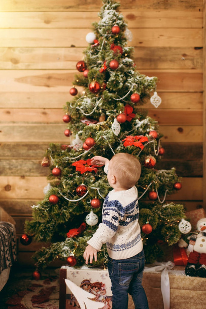 Happy cute little baby boy dressed in sweater and jeans decorating Christmas tree with toys in wooden room at home. Child with good mood. New Year. Lifestyle, family and holiday 2018 concept - Photo, image