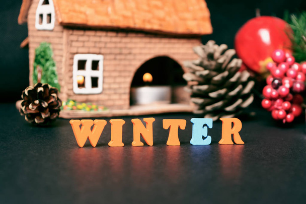 Der Winter ist da. Der Boden ist mit farbigen Buchstaben auf einem schwarzen Tisch auf dem Hintergrund der Zapfen, einem festlichen Kranz aus Weihnachtsbäumen und einem Lehmhaus mit Kerze ausgekleidet.. - Foto, Bild