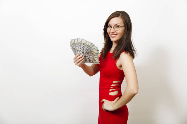 Jolie caucasienne jeune femme d'affaires heureuse en robe rouge et lunettes tenant billets d'argent sur fond blanc. Belle fille avec argent regardant caméra isolé
. - Photo, image