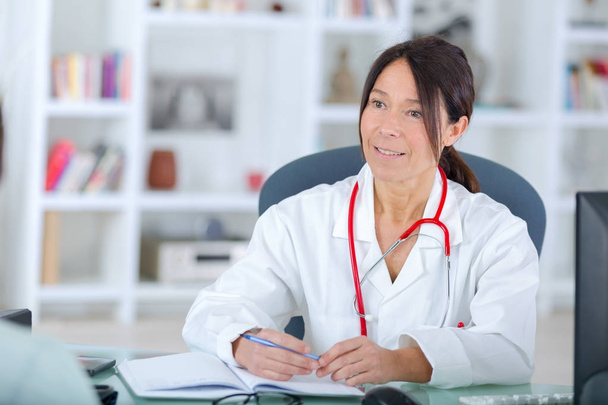 Όμορφη νεαρή χαμογελαστή γυναίκα γιατρός κάθεται στο γραφείο - Φωτογραφία, εικόνα