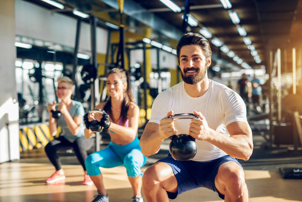 attractive handsome bearded man holding kettlebell and doing squats in fitness group in gym - Photo, Image
