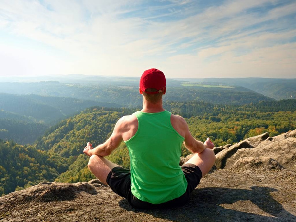 Caminhante corpo magro em singlet verde e shorts pretos sentar em uma rocha, desfrutar de paisagem natural. Vista para o vale da floresta
 - Foto, Imagem