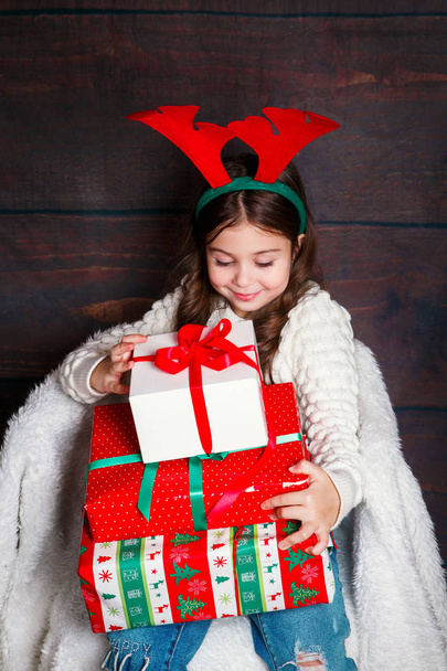 happy little smiling girl with christmas gift boxs..Christmas concept. Smiling funny child in deer horns on wooden background - Foto, Imagen