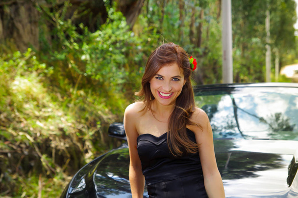 Gros plan d'une belle femme portant une robe noire et une fleur rouge dans sa tête et posant devant une voiture noire de luxe lors d'un road trip, la voiture debout sur les lignes de touche, dans une nature floue
 - Photo, image