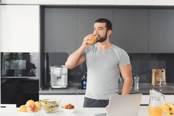 A sporty man stands in the kitchen and drinks orange juice. Next to him on a stack of vegetables and fruits. - Фото, изображение