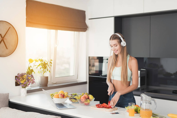 Young athletic girl is standing in the kitchen and is preparing a salad. She has white headphones on her head. - Φωτογραφία, εικόνα