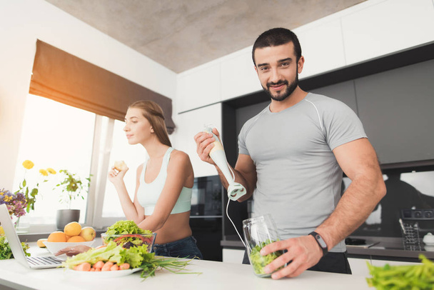 Young sports couple is preparing in the morning in the kitchen. A man is making a green vegetable cocktail. - Φωτογραφία, εικόνα