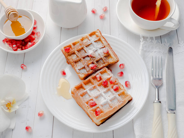 Delicious mouth-watering Viennese waffles with honey and pomegranate seeds on a white plate, light wooden background - Φωτογραφία, εικόνα