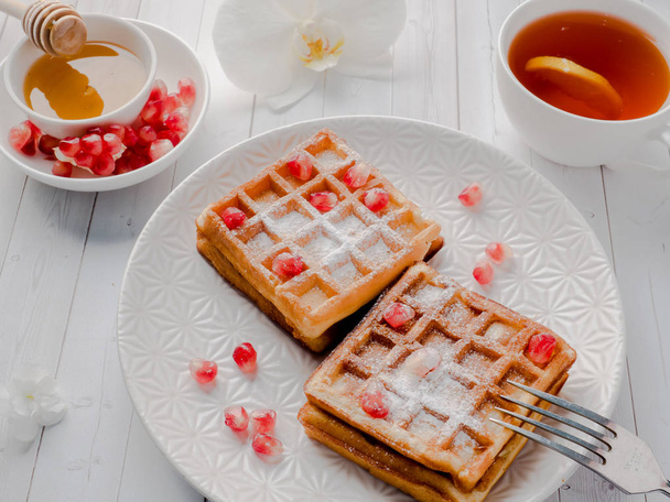 Delicious mouth-watering Viennese waffles with honey and pomegranate seeds on a white plate, light wooden background - Fotó, kép