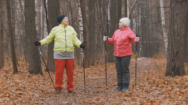 Дві літні жінки в осінньому парку роблять підігрів перед скандинавською ходою
 - Фото, зображення