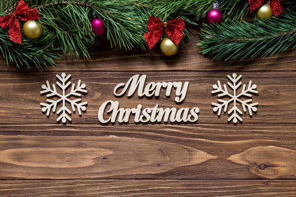 Merry Christmas on the center of the wooden background with pine tree branches on the top of the screen and two beautiful snowflakes - Photo, Image