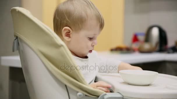 Kicsi fiú ülő-ban a gyerekülés, a konyhában, és a lemezeket nagyon finom étel. Mosolyog és játszik. - Felvétel, videó