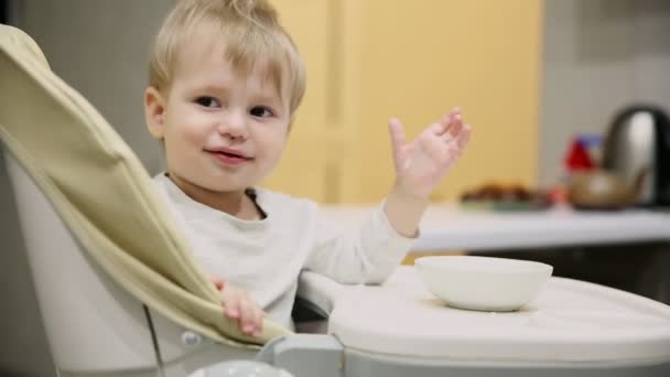 Little boy sitting in the child seat in the kitchen and from the plates is very tasty food. Smiles and plays. - Footage, Video