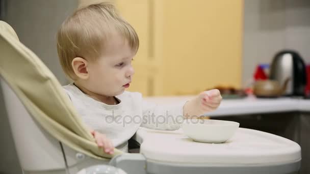 Kleine jongen zitten in het kinderzitje in de keuken en van de platen is zeer lekker eten. Glimlach en toneelstukken. - Video