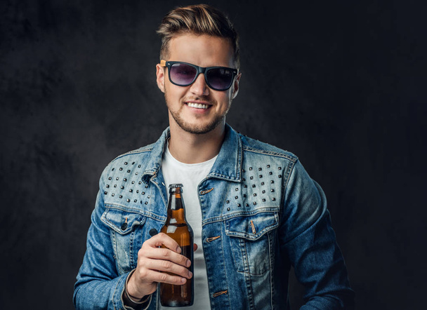 Positive blond male dressed in a denim jacket and sunglasses holds a bottle with craft beer. - Фото, изображение